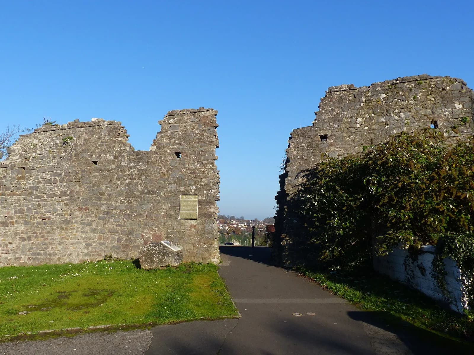 A gap in a stone wall reveals a path leading towards distant buildings under a clear blue sky.